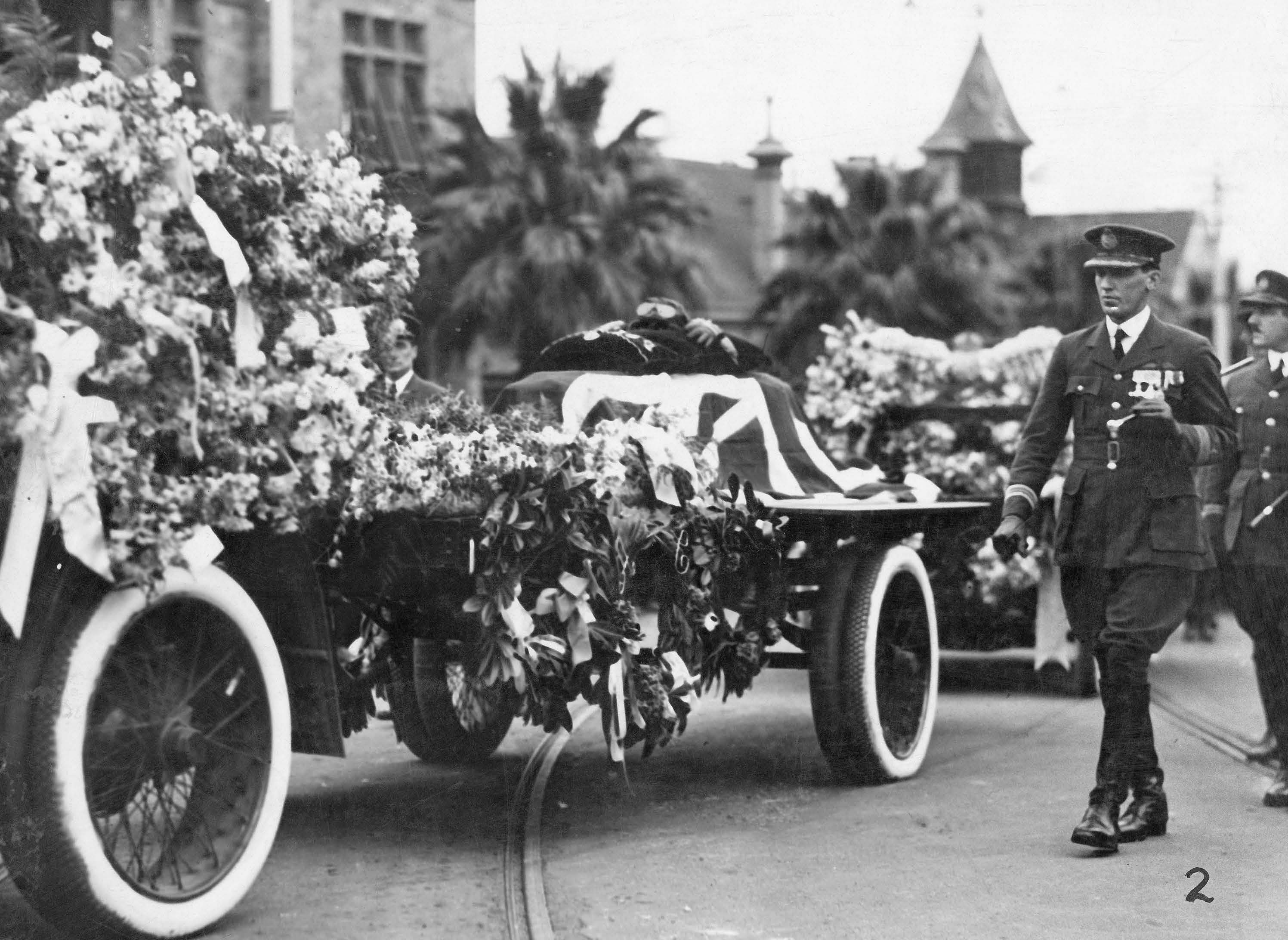The flower laden bier with Ross’ goggles and gloves on his flag draped coffin passing the Children’s Hospital. SLSA: B 876