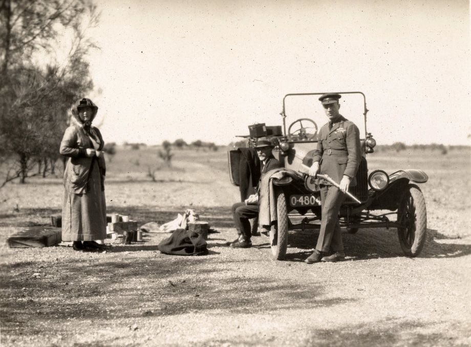 Jessie Smith, with her husband Andrew Smith, and son Ross Smith, enjoying a family picnic at Mutooroo Station, South Australia. Ross is leaning against a car, and is wearing his Air Force uniform and holding a rifle, 1920. SLSA: PRG 18/2/14