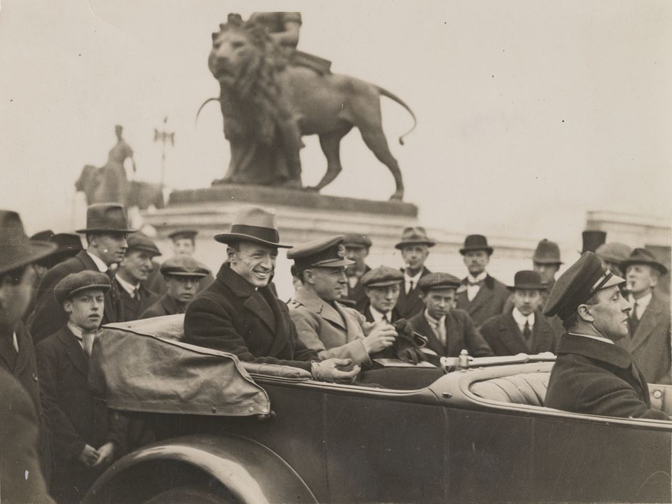 Ross and Keith Smith in a car outside Buckingham Palace, after Keith received his knighthood as a member of the Order of the British Empire (K.B.E.) from King George V, in recognition of the England-Australia flight. A crowd of well-wishers is gathered around the car. SLSA: PRG 18/10/4