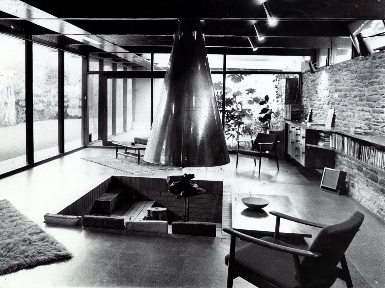 Interior view of a house on Waterfall Gully Road, with an internal fire pit designed by John S. Chappel Pty Ltd, architect. Showing the living room with picture windows and a feature stone wall, fire pit in the centre, with cushions for seating inside and large metal hanging flue, 22 June 1969. SLSA: BRG 346/29/181/2